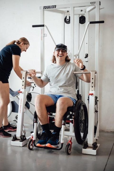 A young man in a wheelchair, smiling to the camera about to use exercise equipment in a disability rehabilitation centre.