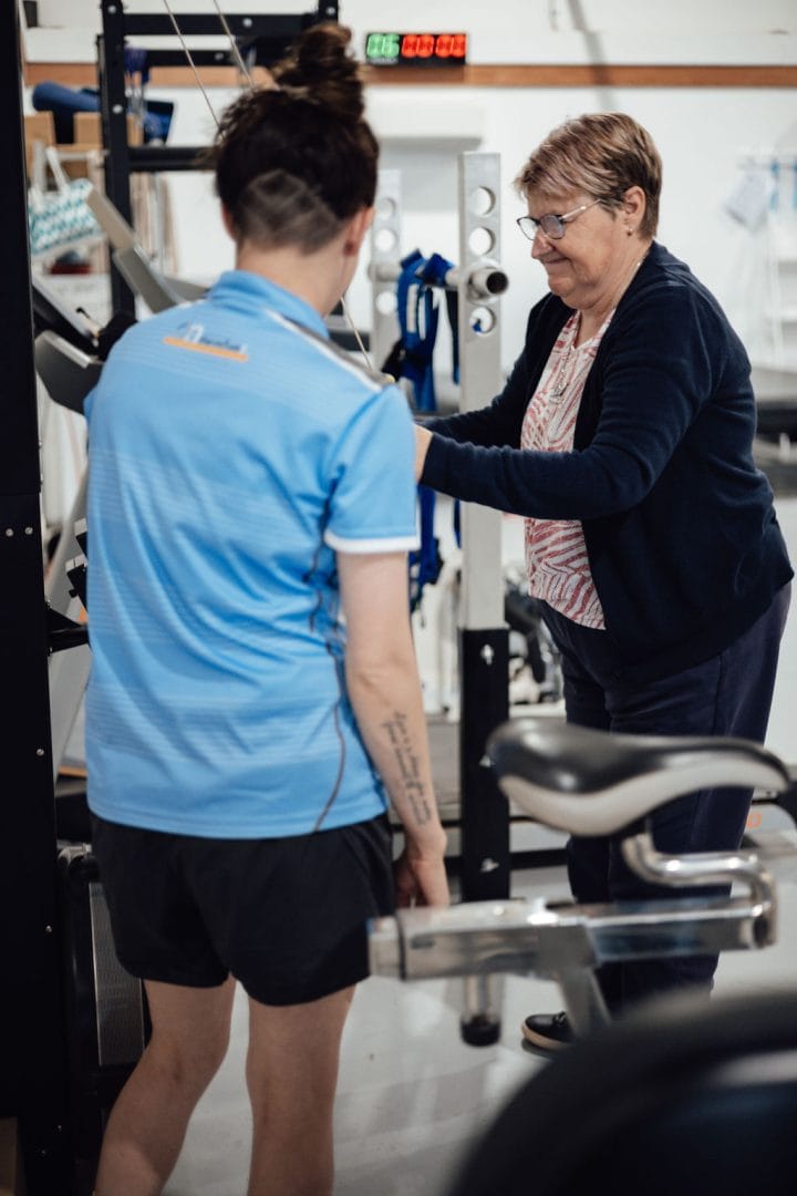 An exercise therapist in a blue shirt is guiding an older woman to use gym equipment.