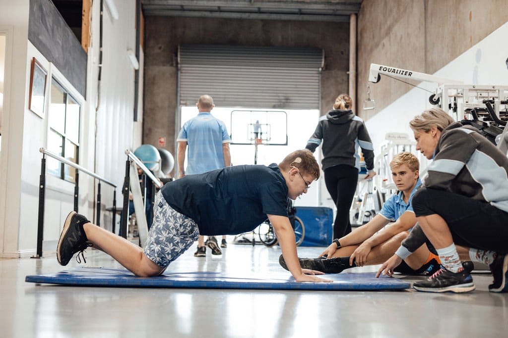 A young disabled boy doing pushups in a disability rehabilitation centre with two exercise physiotherapists encouraging him.