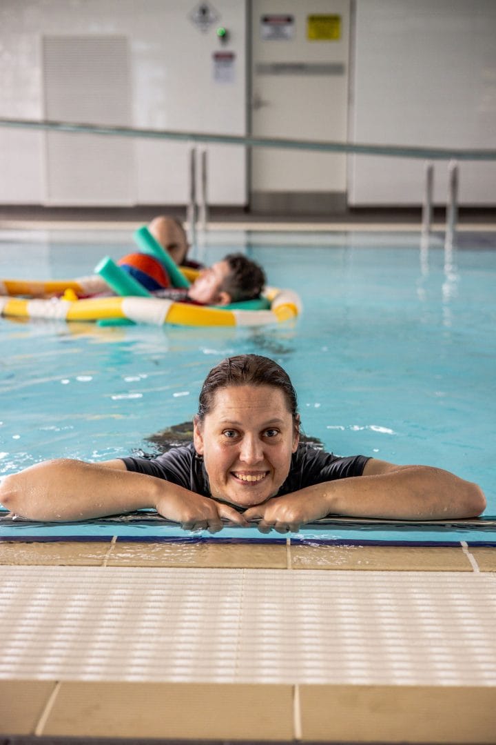 A woman in a hydrotherapy pool smiling at the camera with an exercise therapist the background with a disabled participant on a float.